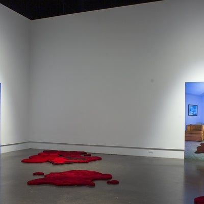 Art installation in a gallery with two large photographs of room interiors, one showing a woman's legs on the floor and other an orange armchair and red rug. On the gallery floor are several irregularly shaped red floor rugs that resemble pools of blood.