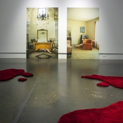 Art installation in a gallery with two large photographs of room interiors, one showing a white woman’s body under a sofa cushions and in the second she is lying on the linoleum floor between an old orange armchair and a stereo cabinet.