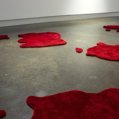 Several irregularly shaped red floor rugs, that resemble pools of blood, are scattered across a concrete floor.