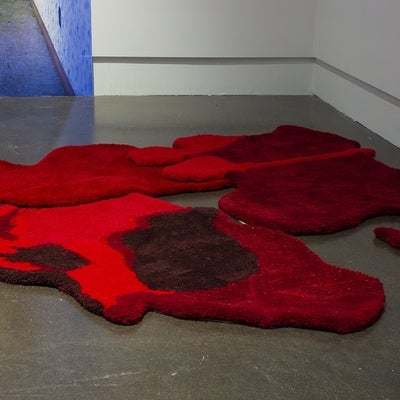 An art gallery with several irregularly shaped red floor rugs, that resemble pools of blood, on a concrete floor. Behind them is a corner of a photograph showing a room with a linoleum floor and a woman's feet.