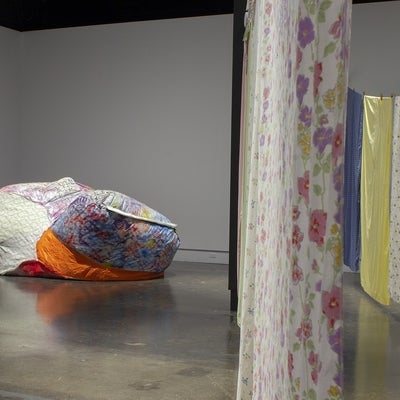 View of an art exhibition with multi-coloured and patterned bedsheets hung on what resemble clotheslines and a large, irregularly shaped, inflated sculpture made from various bright fabrics. 