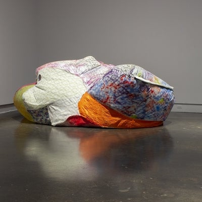 A large, irregularly shaped, inflated sculpture made from various bright fabrics on a gallery floor. 