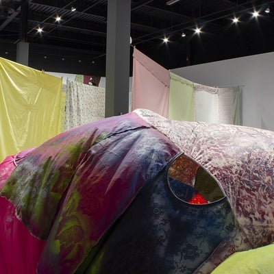 View of an art exhibition with a large, irregularly shaped, inflated sculpture made from various bright fabrics in front of multi-coloured and patterned bedsheets hung on what resemble clotheslines. 