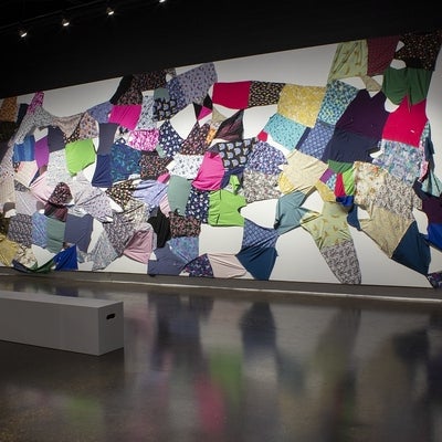 An artwork with multi-coloured and patterned caregiver scrubs collaged together in a quilt-like pattern and stretched to cover a wall.