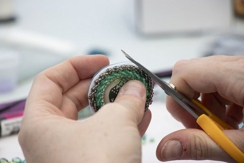 Close-up of person's hands while they use scissors to trim a beaded medallion.
