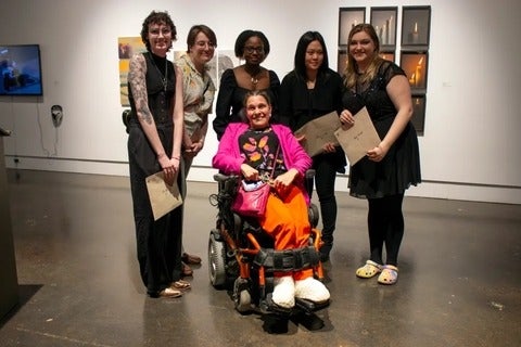 In an art gallery, five people holding envelopes gather around a sixth person in a wheelchair.