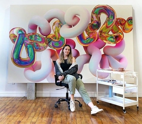 Person sits casually in a rolling office chair in front of a large painting of rainbow coloured tubular shapes.