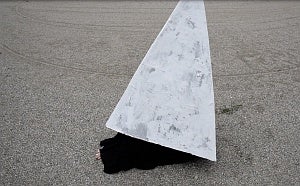 In a gravel lot, a figure, wrapping in black, sits on the ground inside a light grey pyramidic form with only their toes showing
