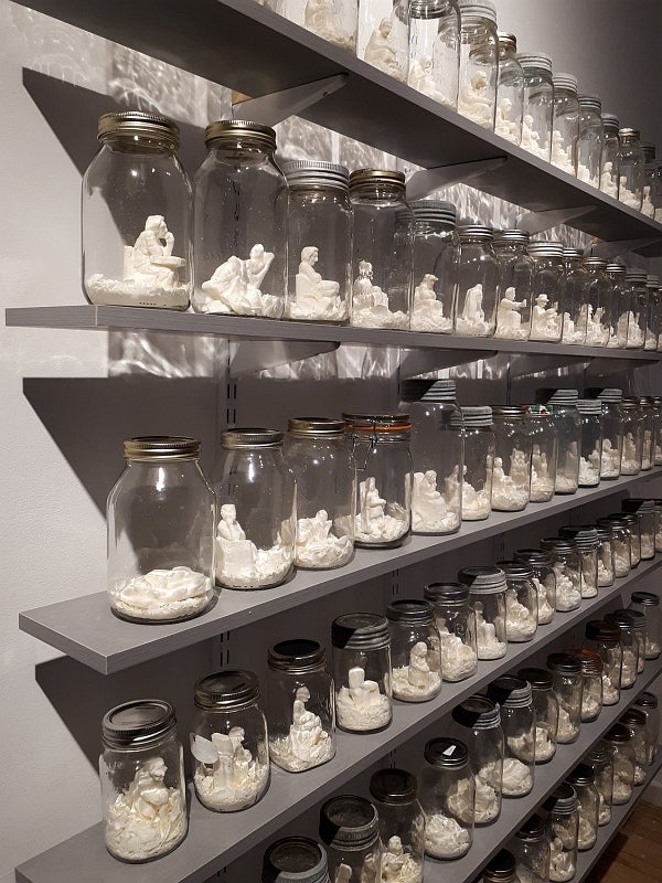 Six shelves filled with mason jars containing sculptures of white seated figures.