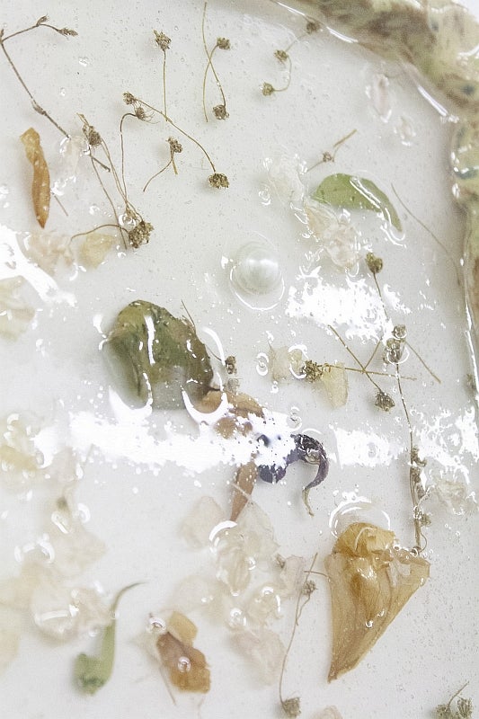 Detail of artwork with flower petals, leaves and pearl embedded within a glossy resin on a white surface.