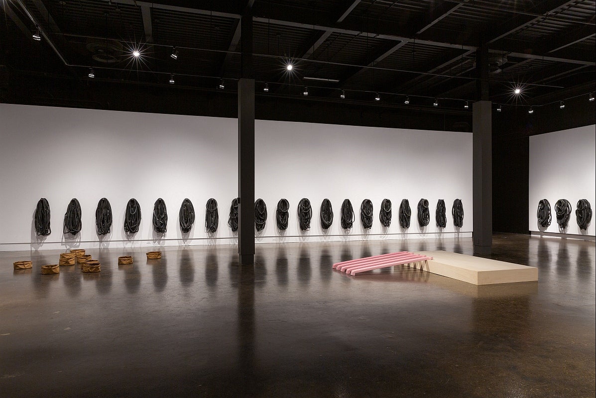 Artwork in a gallery. On walls, multiple groups of bike tubes. On floor, series of large round paperbags and pink cast eaverstro