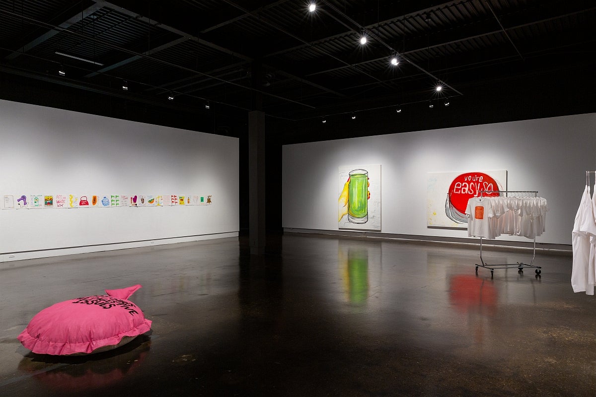 Exhibit with a series of colourful drawings pinned to the wall, 2 large paintings, t-shirts and a large pink cushion on floor
