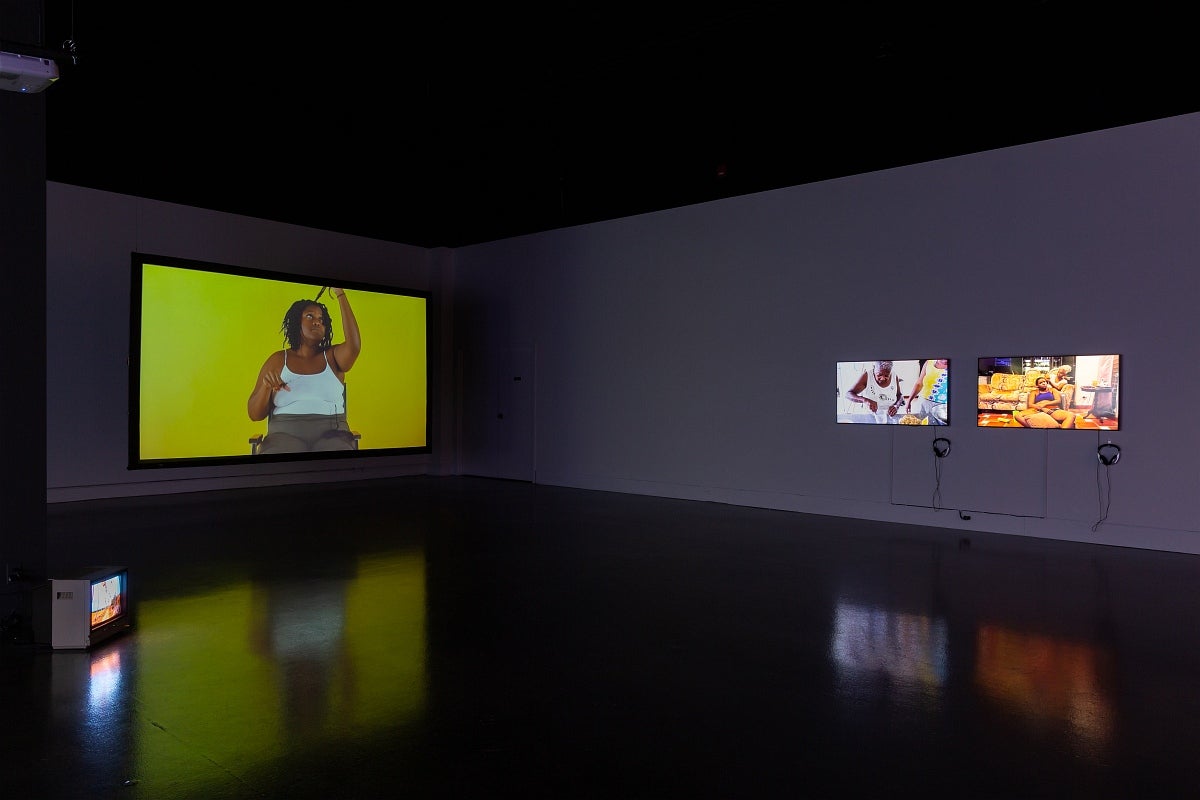  Video exhibition in dark gallery. Video at left show person in a chair cutting sections of their hair, at right videos show peo
