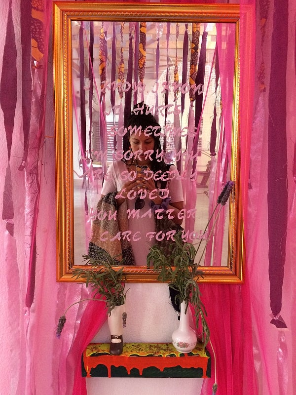 Person takes a photograph in front of a mirror while surrounded by hanging red and pink ribbons, text on mirror reads I know, I
