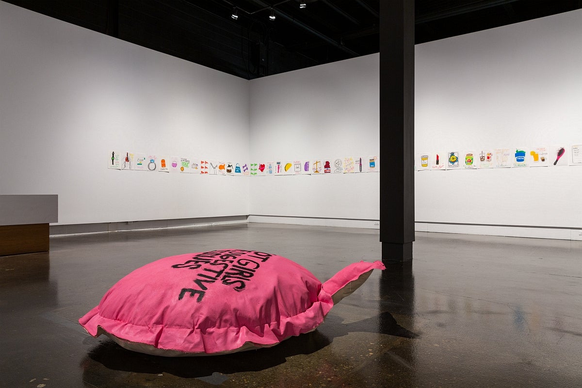 Art exhibit with a series of colourful drawings pinned to the wall in a row and a large pink cushion on the floor.