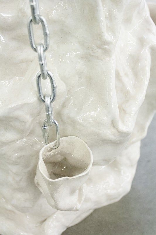 White ceramic sculpture of a sack-like vessel filled with water and hanging from a chain attached to larger ceramic vessel