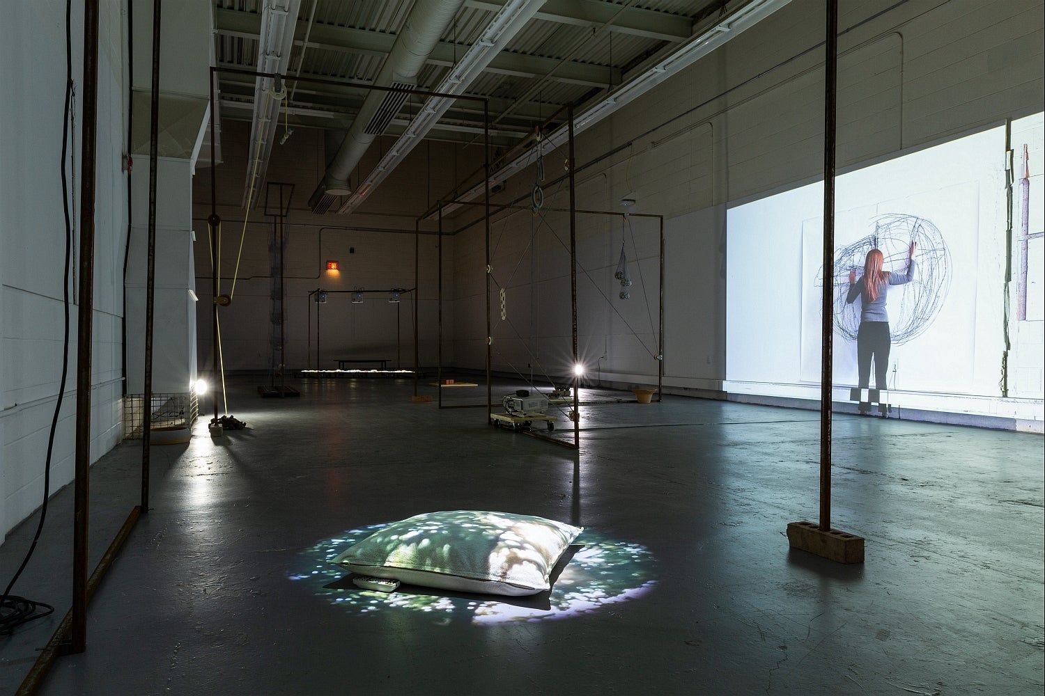 Dark, industrial space with art installation. Projectors light objects on the concrete floor and a video of a person drawing.