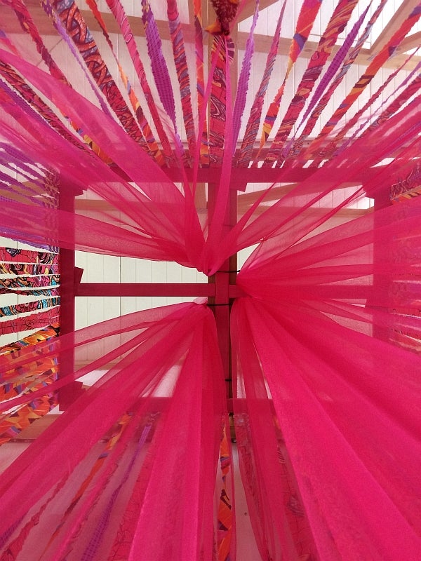 View inside a fabric sculpture, looking up at red and patterned ribbons hanging down from a wood frame.