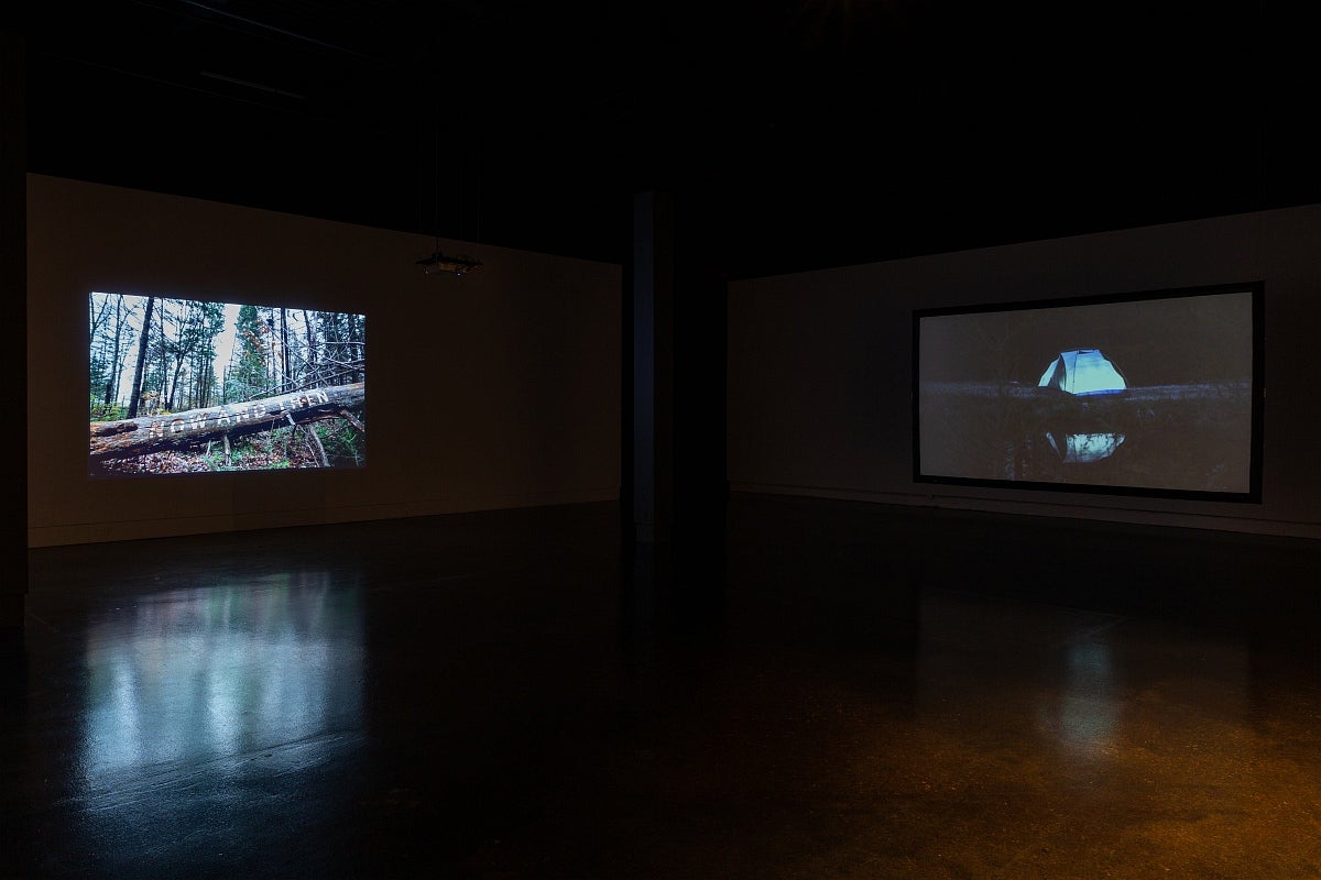 Video exhibition in dark gallery.  Video on left shows person carving letters on a fallen tree, right video of glowing blue tent