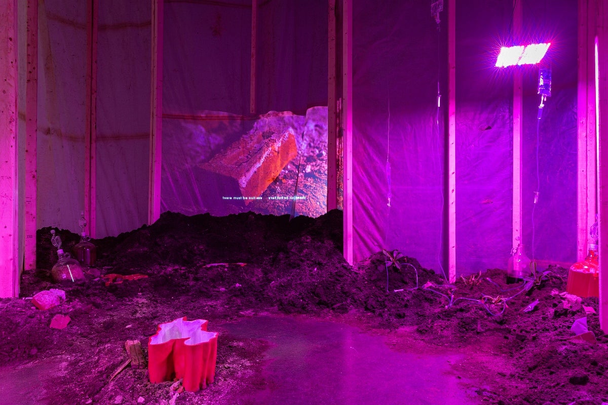Artwork of a room made of wood and plastic sheeting lit magenta, containing mounds of earth and debris, projection of a brick wi