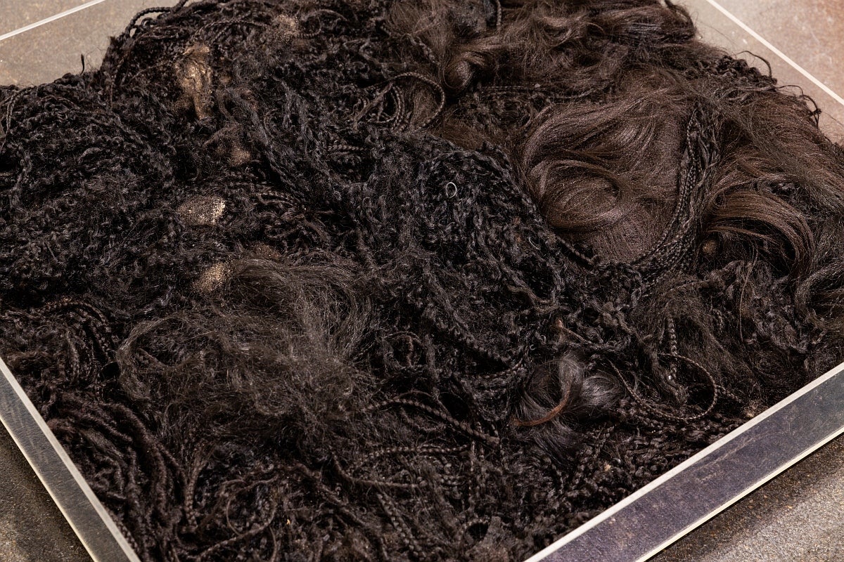 Artwork in gallery. Detail view of long dark hair, much of it in thin braids, filling a low plexiglass box on the floor.
