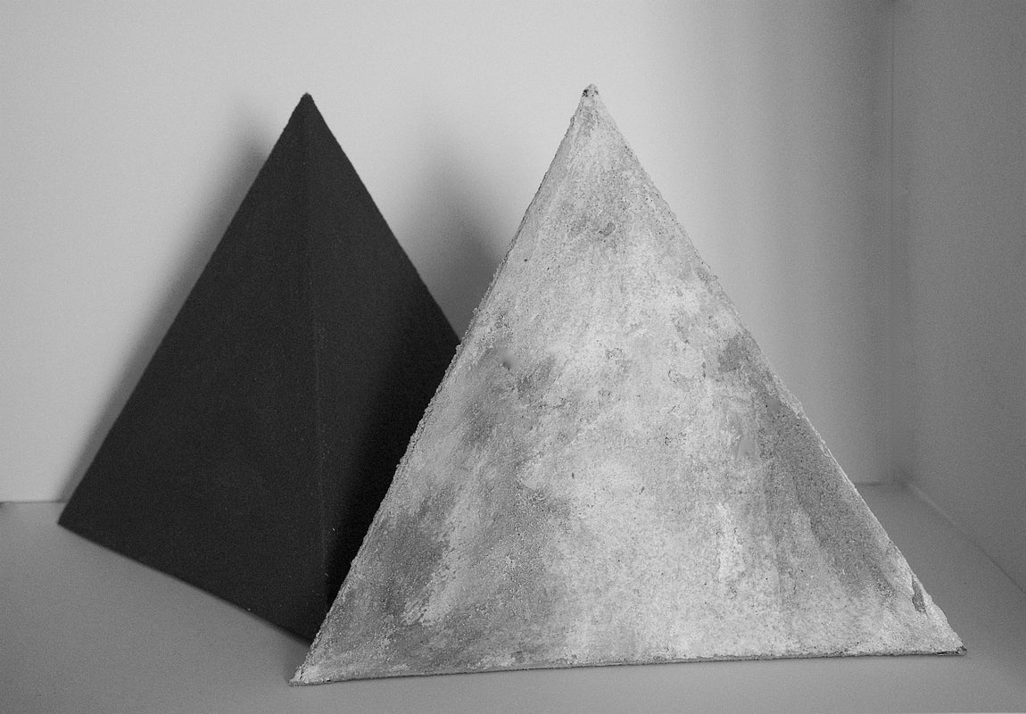 Two small pyramids with rough, irregular surfaces. The pyramid in front is shades of light grey, the one behind is black.