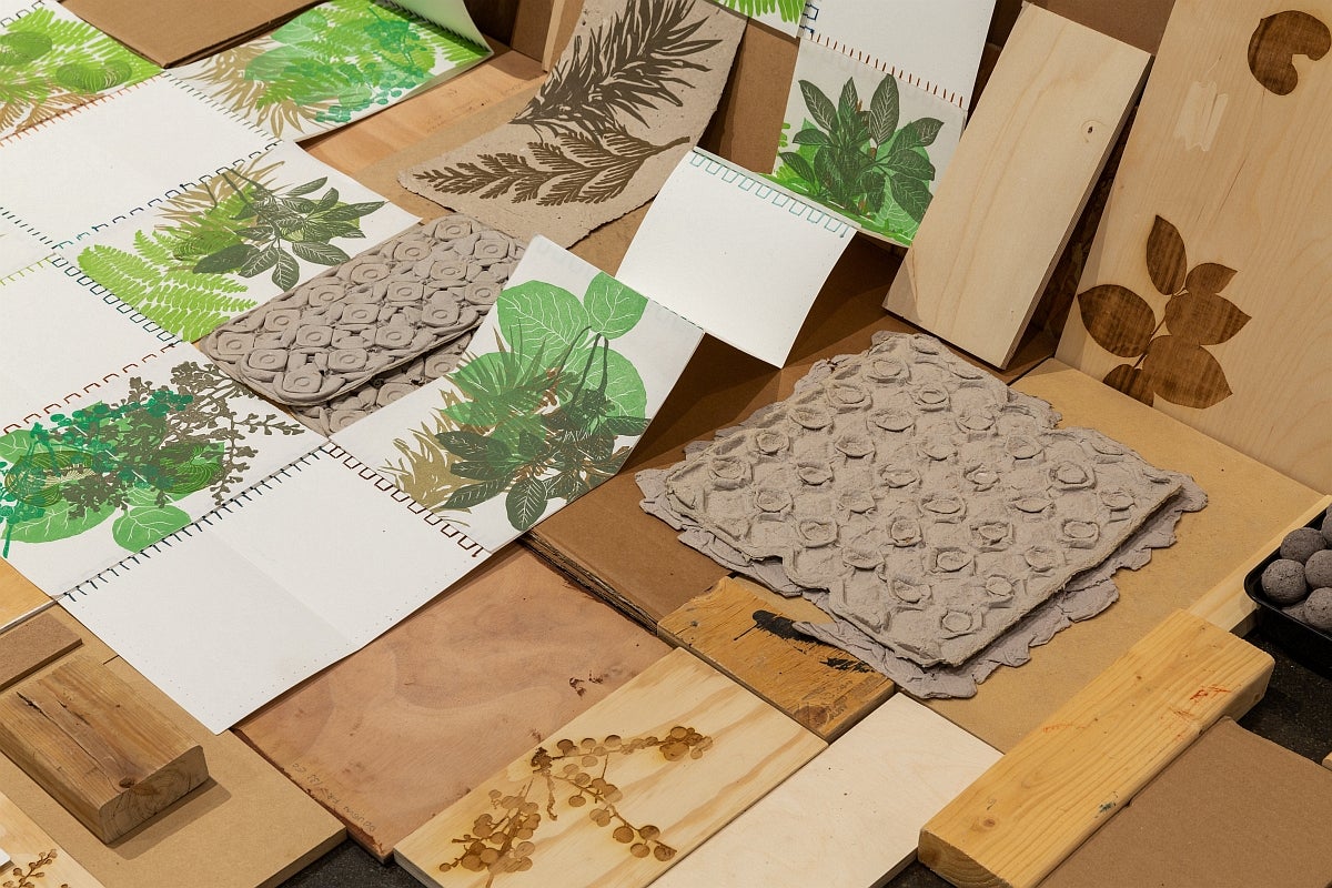 Floor installation of prints of green and brown plants and foliage displayed flat and accordion-style on reclaimed wood.