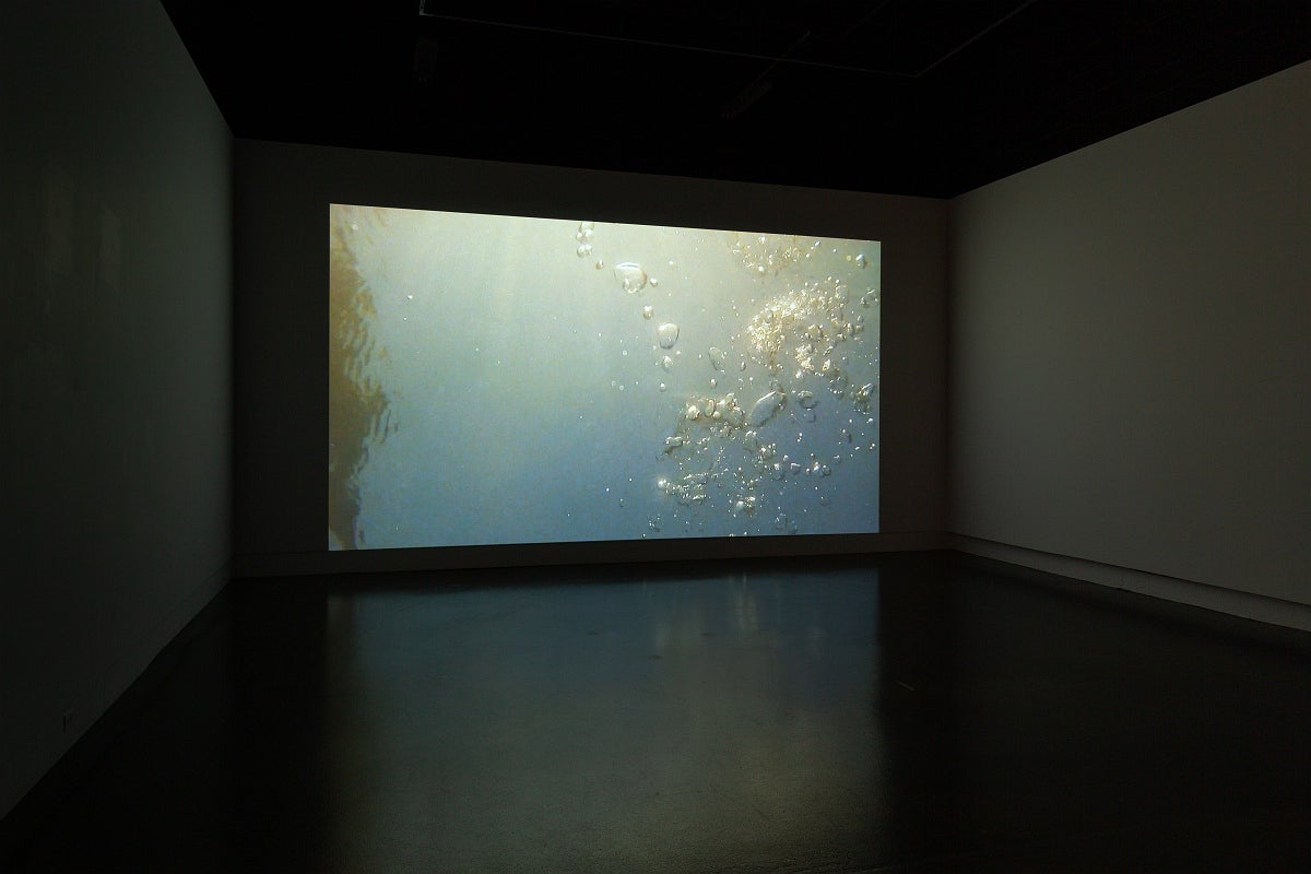 Video in dark gallery. Video show air bubbles rising through water, blue-grey water at bottom which it lit by sun at top