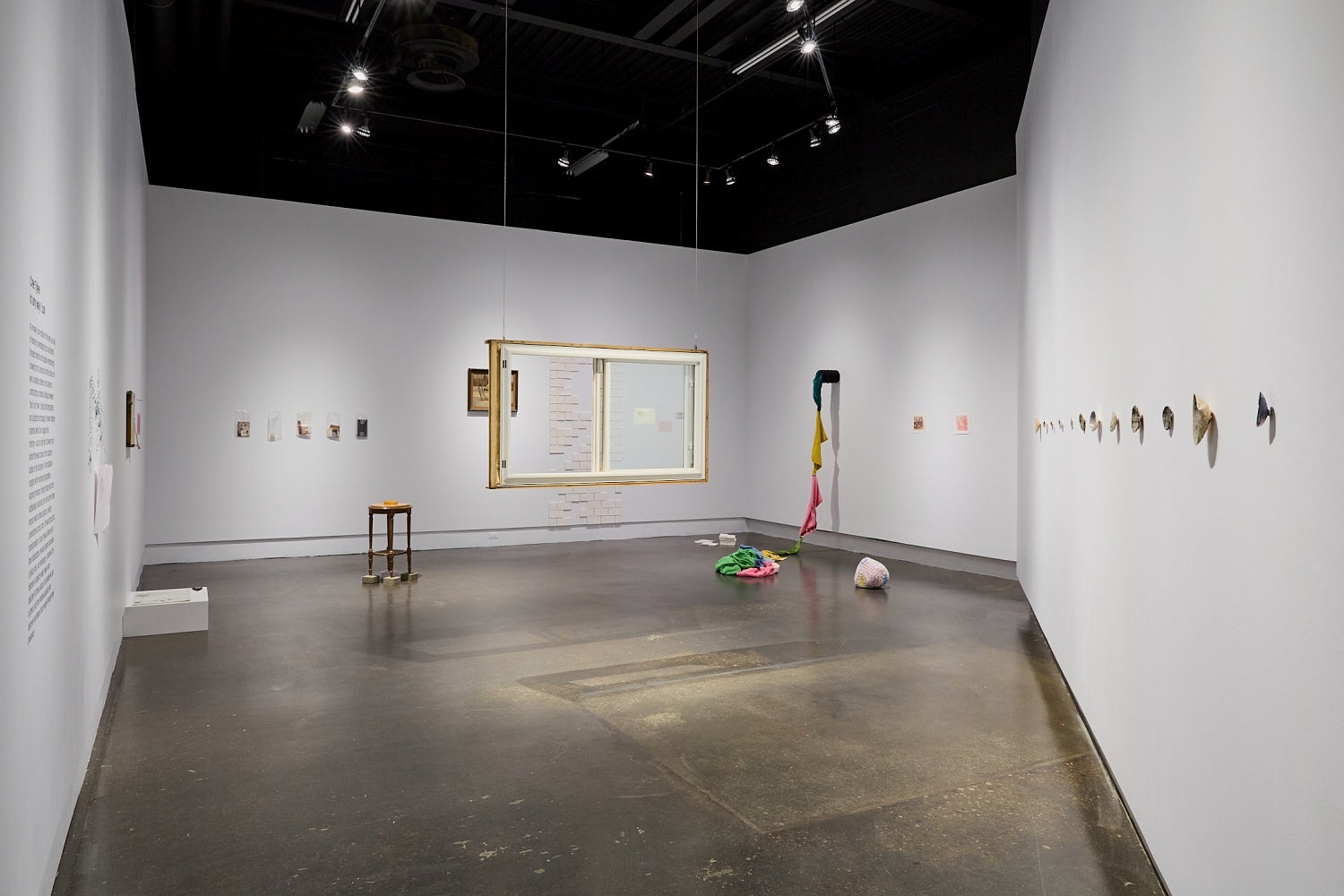 Art gallery installation of multiple ephemera, white ceramic tiles, shells, crocheted items and a salvaged house window hanging in the center of the room.