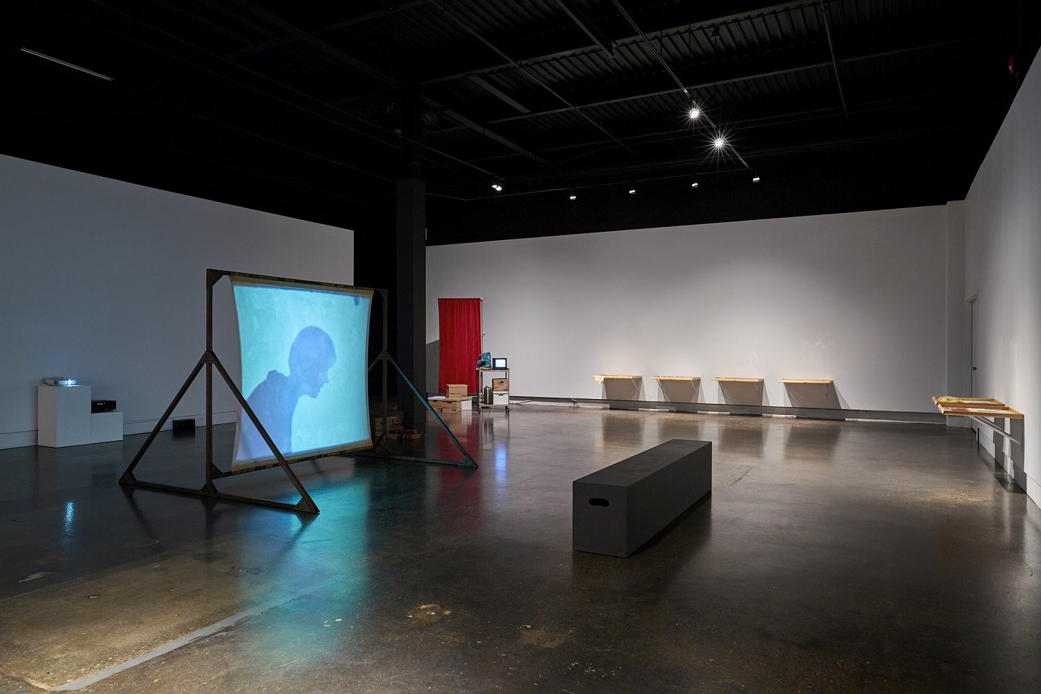 Art gallery installation with videos, one projected on a free-standing screen, wooden drawers attached side by side on a wall and a metal cart and privacy screen in the corner.