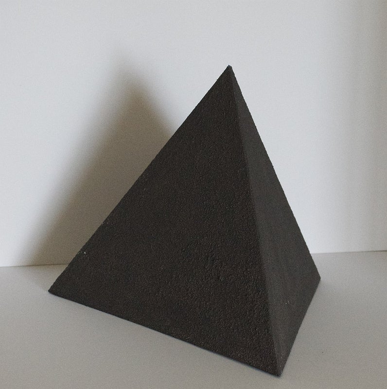 Small, black. pyramid with rough, irregular surfaces in front of a white wall.