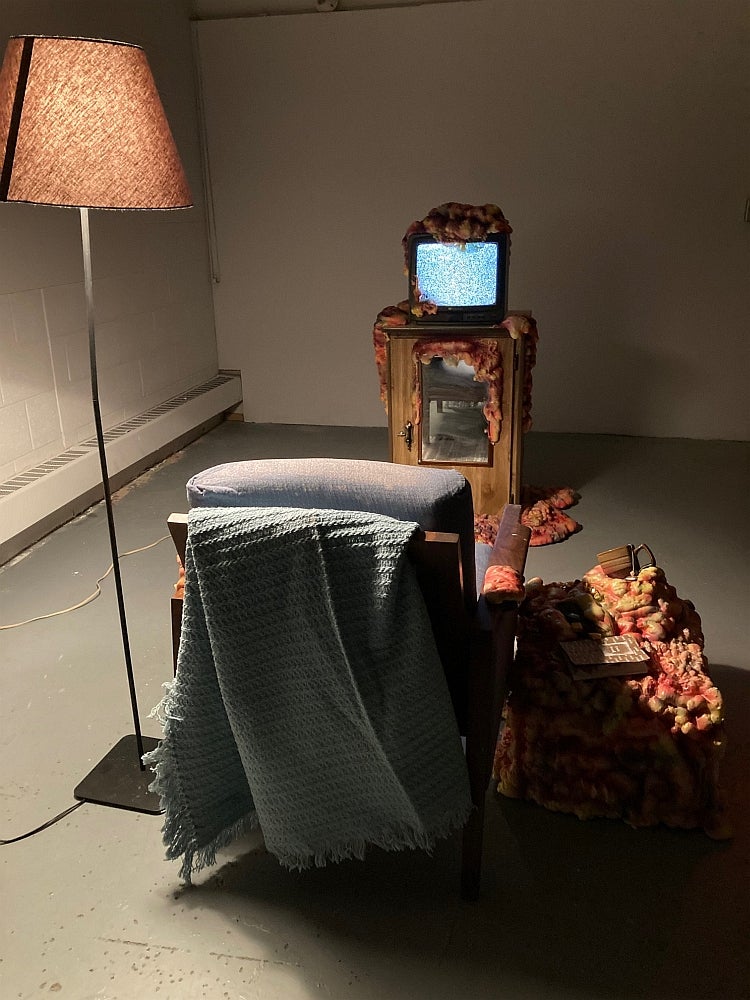 Art installation of an armchair, table, lamp and televison playing static, all topped with spray foam that appears to spread