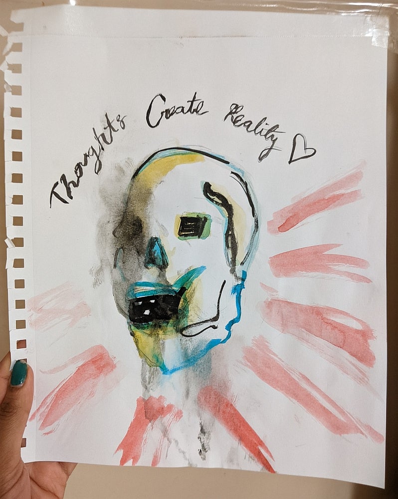 Hand holds a watercolour painting of a skull with the text "thoughts create reality"