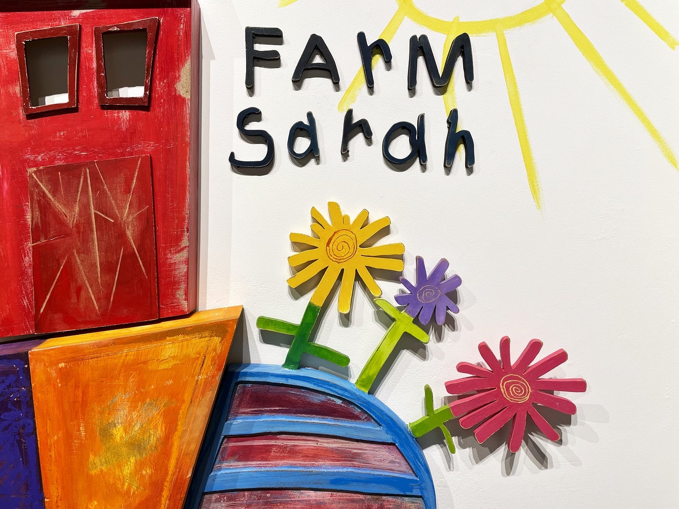 Detail of artwork consisting of painted wooden shapes arranged to form a childlike red barn and flowers, text "Farm Sarah"