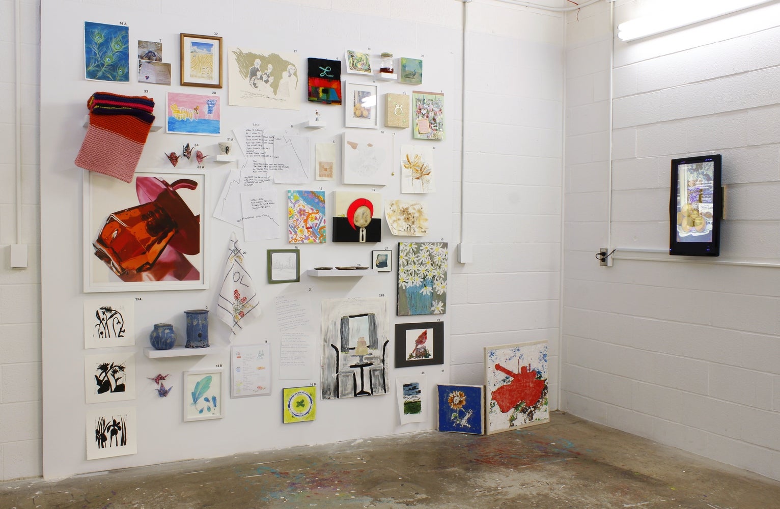 Installation of art in an industrial space. One wall is filled with a variety of works, third wall has small video monitor 