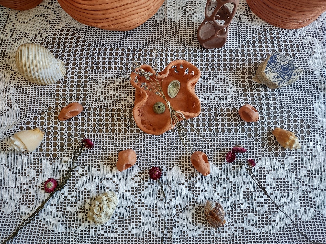 Collection of items including clay vessels, clay skull-like objects, dried flowers and shells sit on the floor on a crocheted t