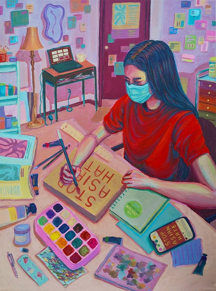 Painting of a girl wearing a face mask sits at a desk painting. Her canvas reads "Stop Asian Hate"
