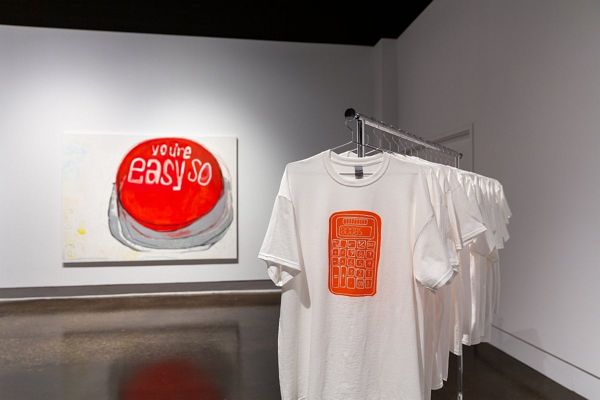 Painting of a red button reading “you’re so easy”, and a rack of white t-shirts with an image of an orange calculator with 80085