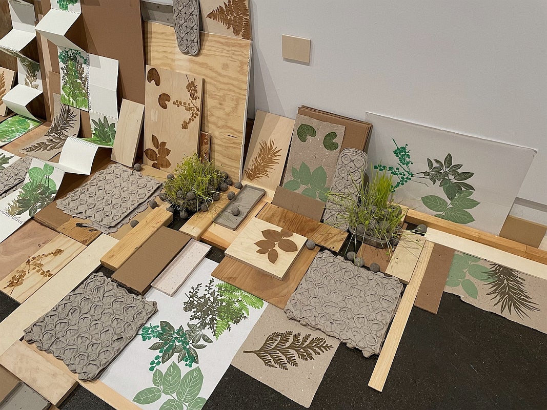 Floor installation of seed bombs, some with sprouting microgreens, and lino-prints of green and brown plants and foliage display
