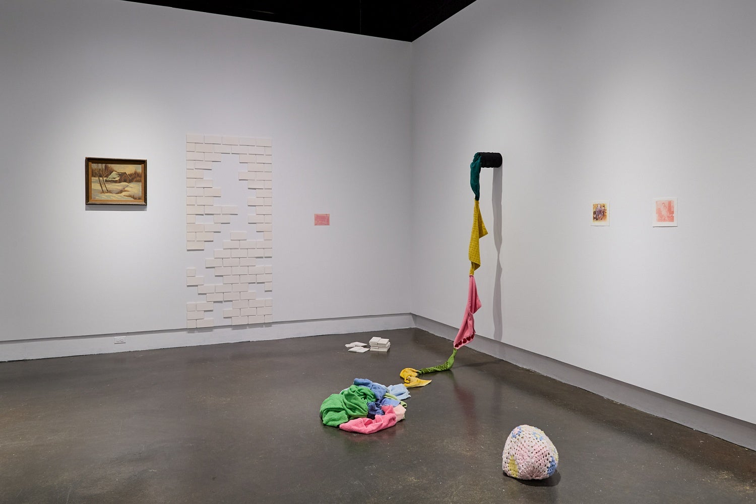 Art gallery installation of brightly coloured terry cloth towels sew together to form a rope, a crochet covered rock, white ceramic tiles, and a vintage landscape painting.