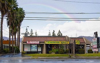 Multiple signs advertising a psychic are displayed on the façade of a suburban mall bordered by palm trees. A rainbow appears in the distance.