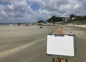 Artist's easle with blank canvas in front of a sandy beach with distant sunbathers.