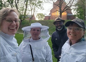 Close-up of four people standing in a green-space with a red brick church behind them.  Three of the people wear white bee-keeping costumes.
