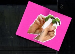 A fuchsia pink paper with a cut out photo of two hands wearing finger puppets on green clawed feet, sits on a black background.