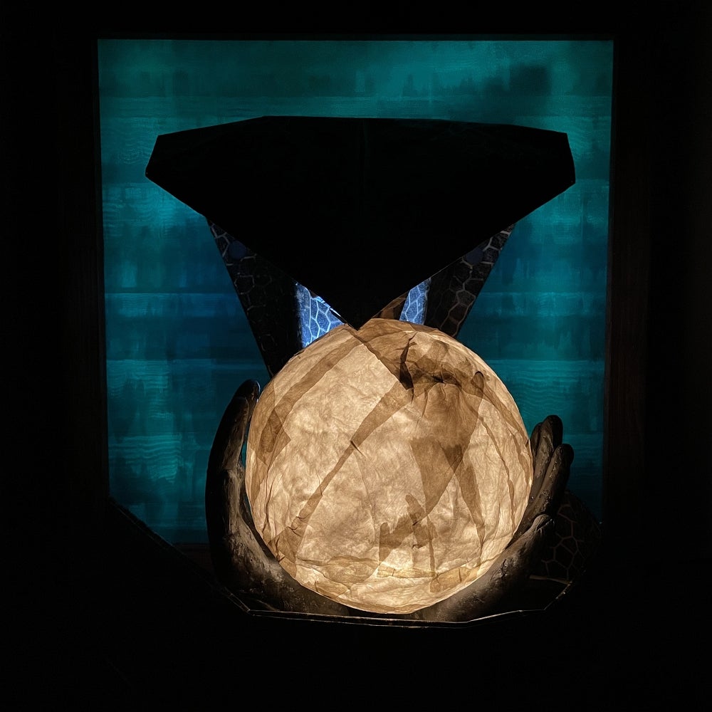 Artwork of glowing papier mache sphere is held in two hands with a paper cobra head above against a pattern teal coloured backgr