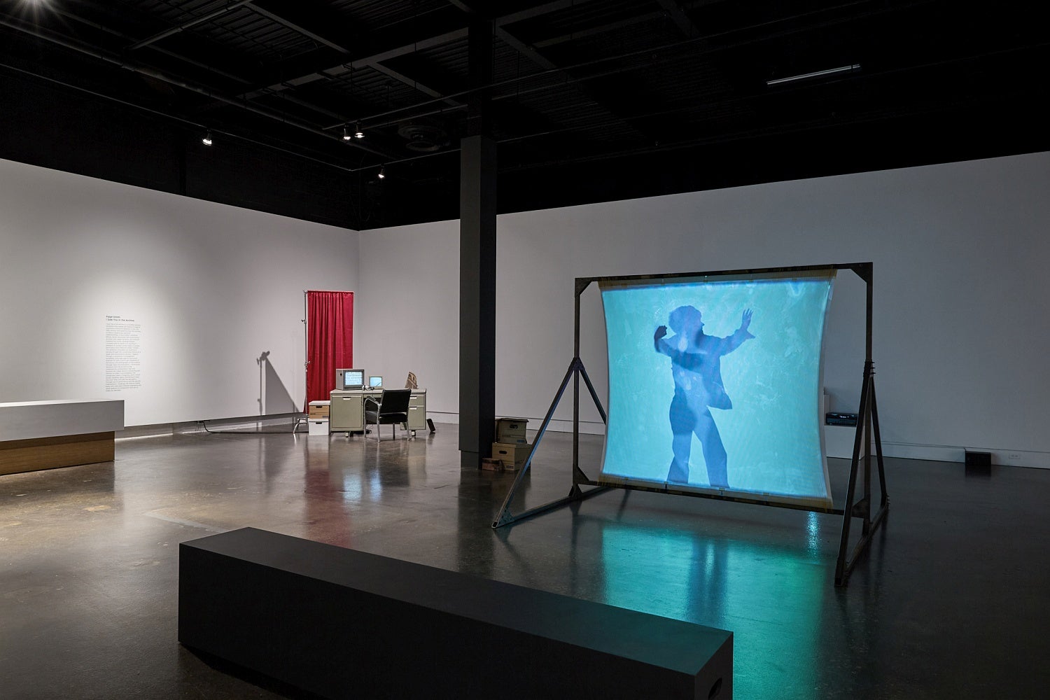 Art gallery with installation of videos, one projected on a free-standing screen, an industrial office desk, chair and privacy screen and multiple bankers boxes.