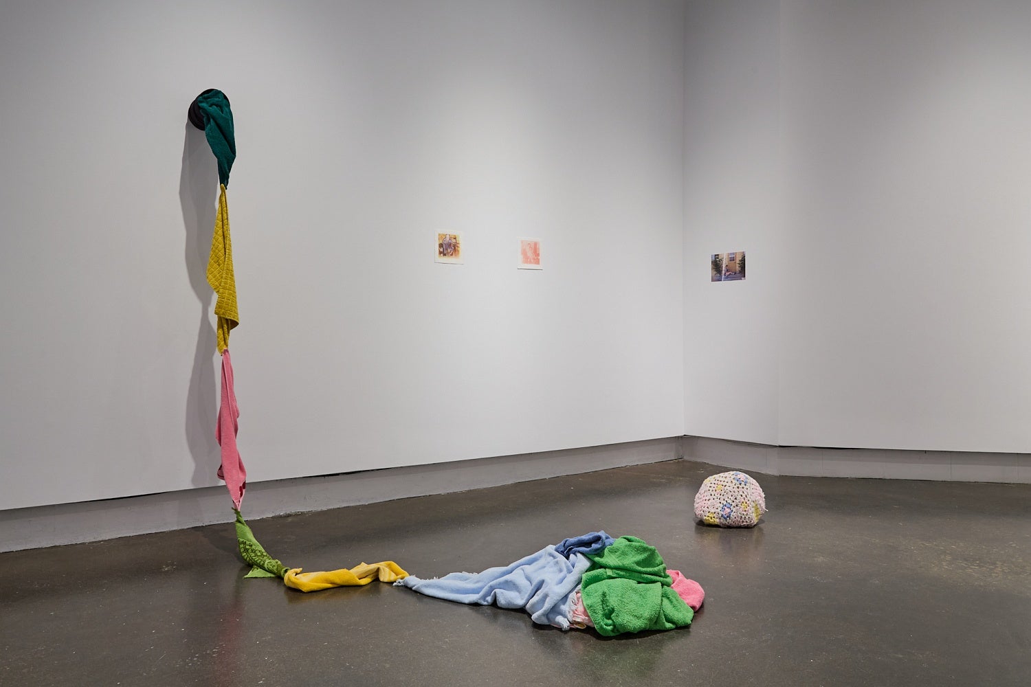Art gallery installation of brightly coloured terry cloth towels sew together to form a rope, a crochet covered rock, and three small photo reproductions.