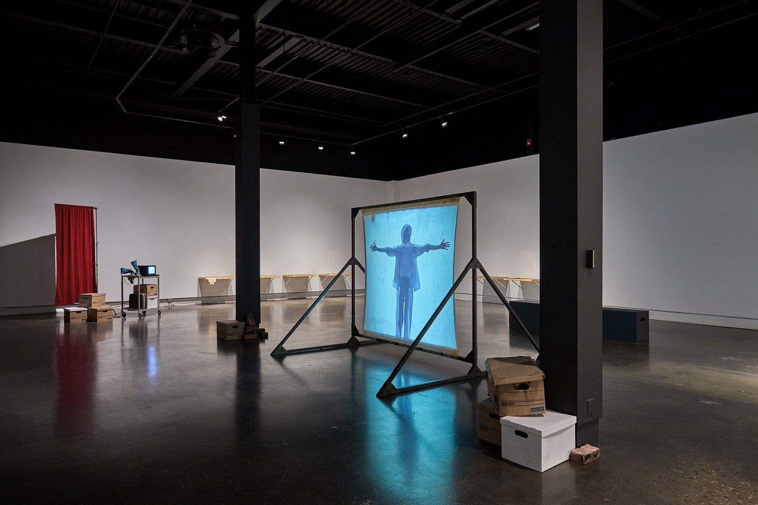 Art gallery installation with videos, one projected on a free-standing screen, wooden drawers attached side by side on a wall and a metal cart and privacy screen in the corner.