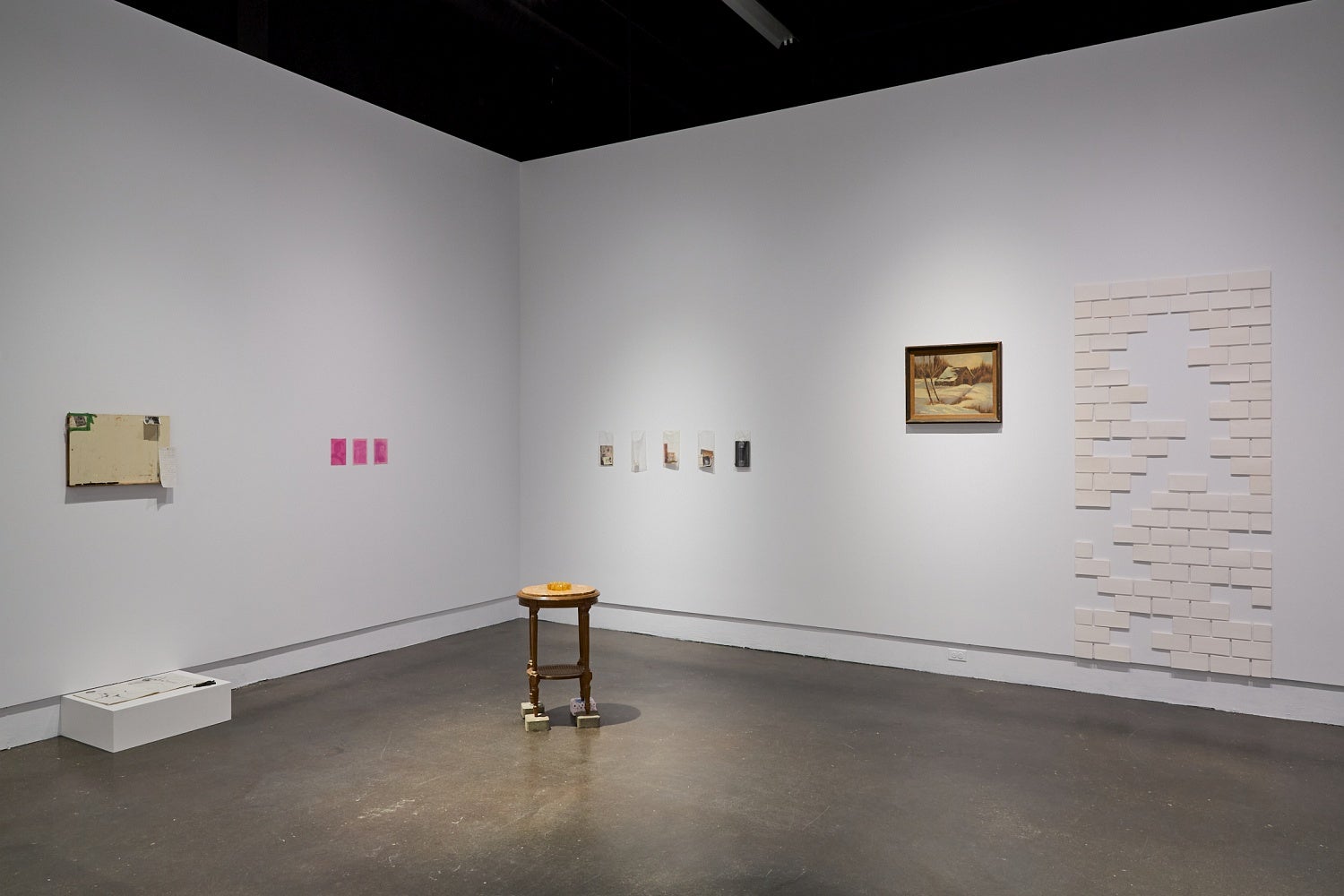 Art gallery installation of vintage ephemera, and marble topped table standing on bricks, white ceramic tiles, and a vintage landscape painting.