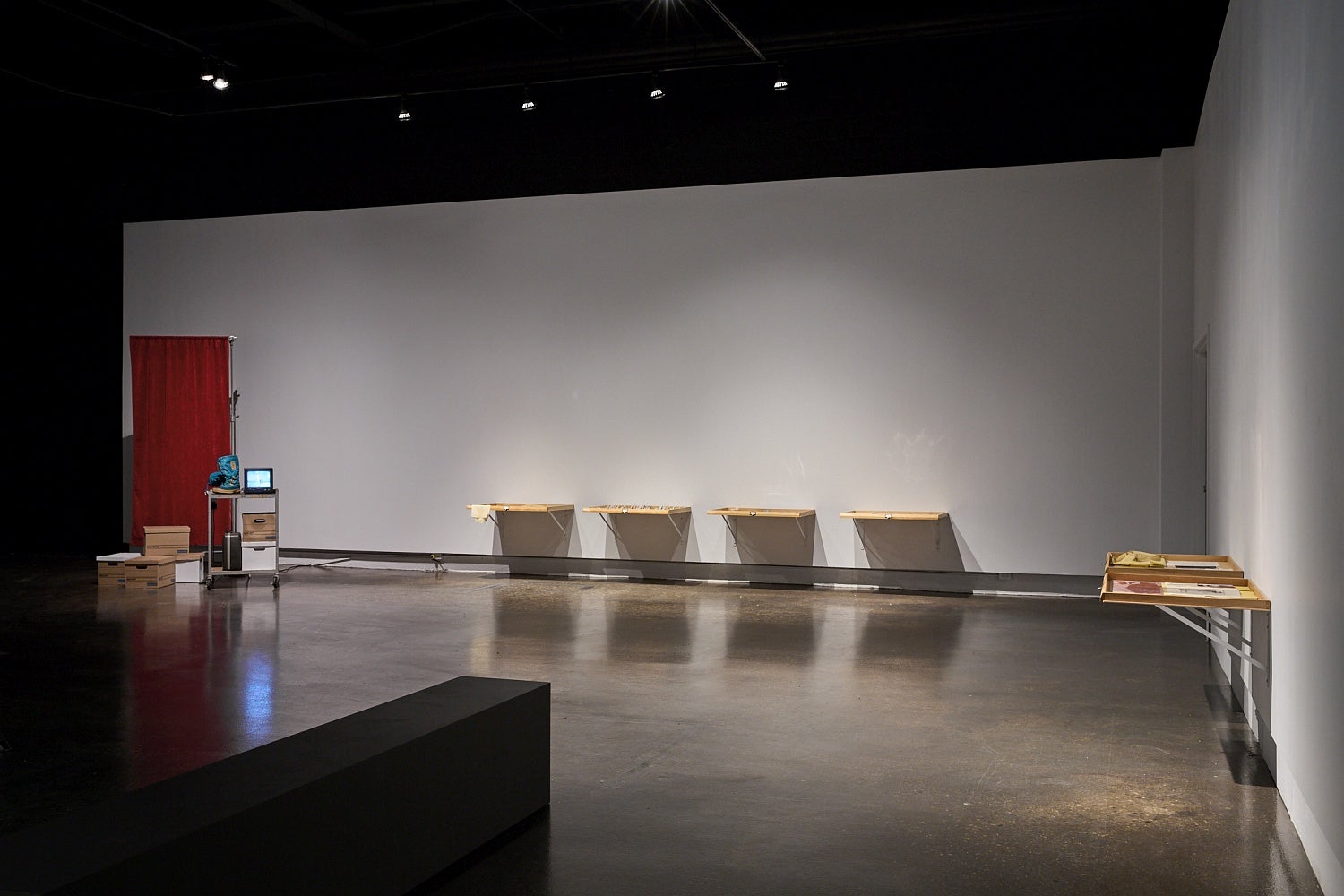 Art gallery with wooden drawers attached side by side on a wall and a metal cart holding a small television, boots and bankers boxes sits in front of a privacy screen.